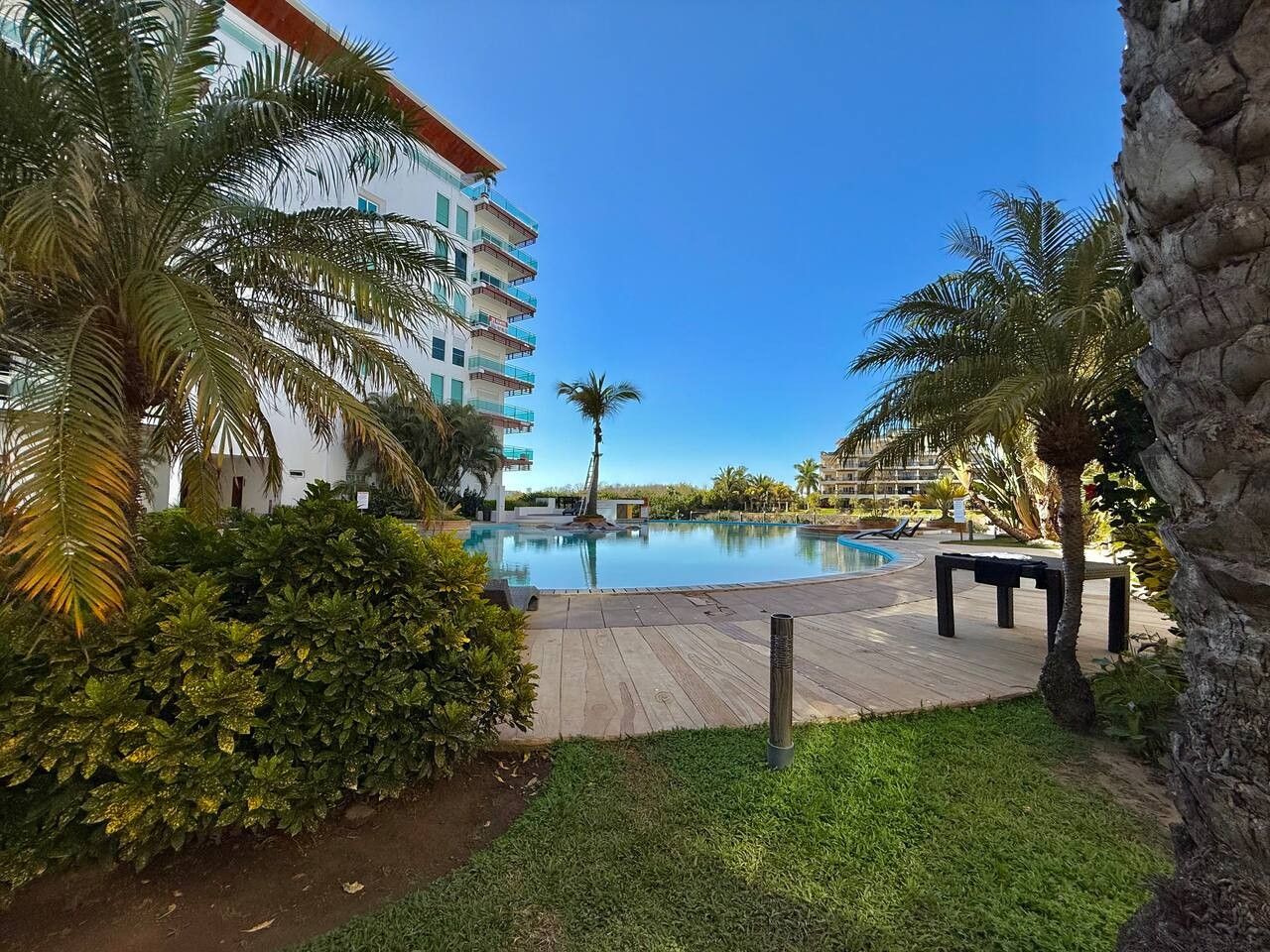 Area piscina di un resort con terrazza in legno, palme e un edificio sullo sfondo.