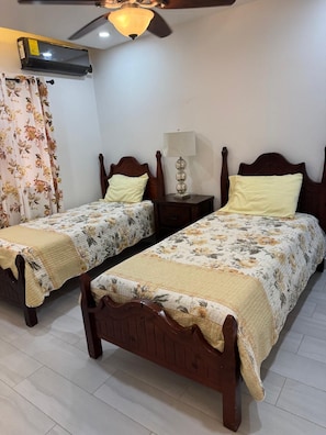 Une chambre avec deux lits en bois, un ventilateur de plafond, un appareil de climatisation, une table de chevet avec une lampe et un rideau à motifs floraux.