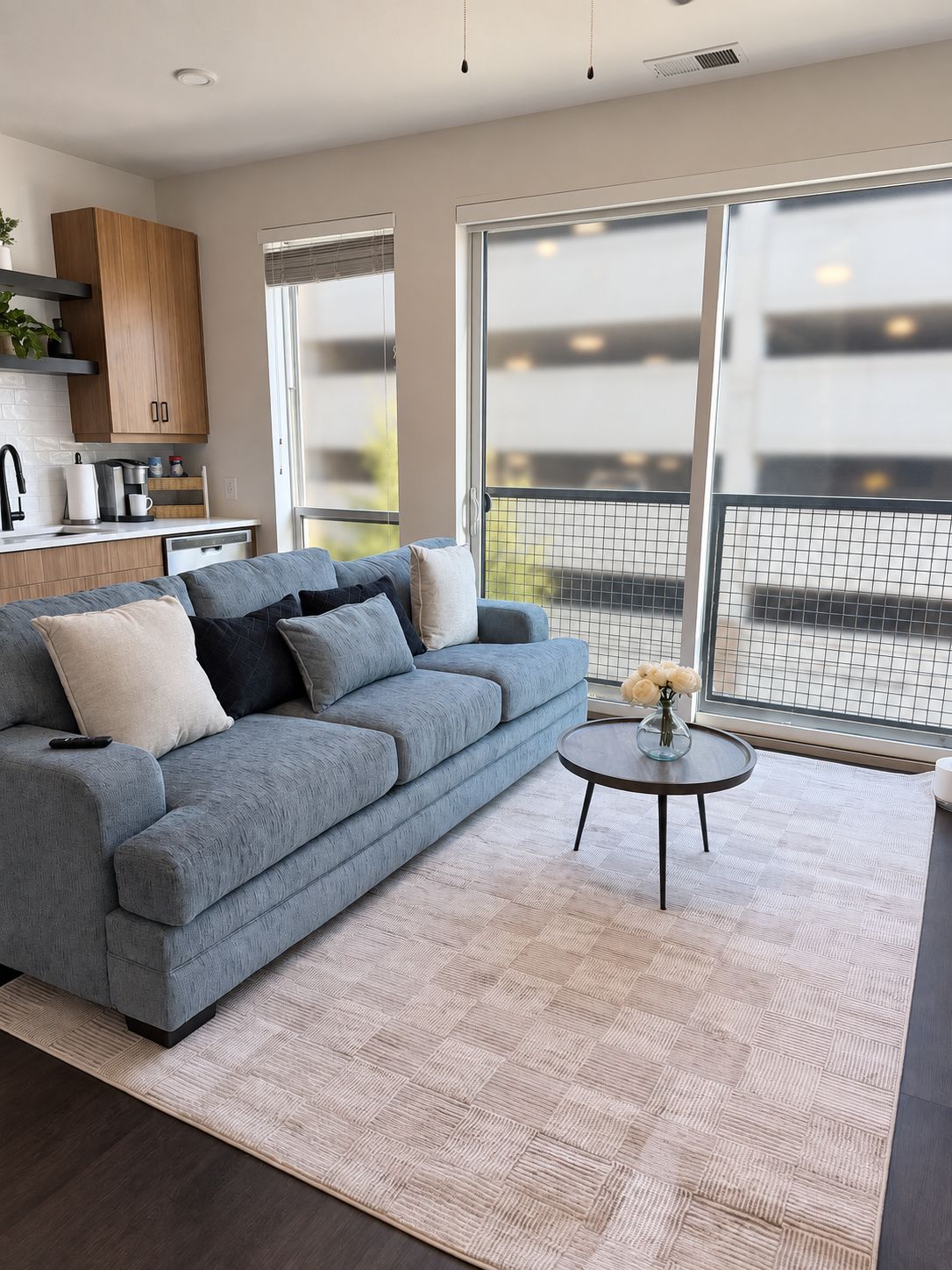 Een moderne woonkamer met een grijze bank, een klein rond tafeltje en een keuken met houten kastjes.