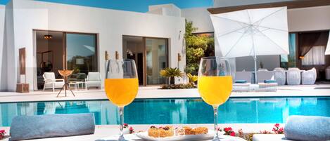 Escena junto a la piscina con dos vasos de jugo de naranja sobre una mesa, una bandeja de postres y un área lounge moderna al aire libre.