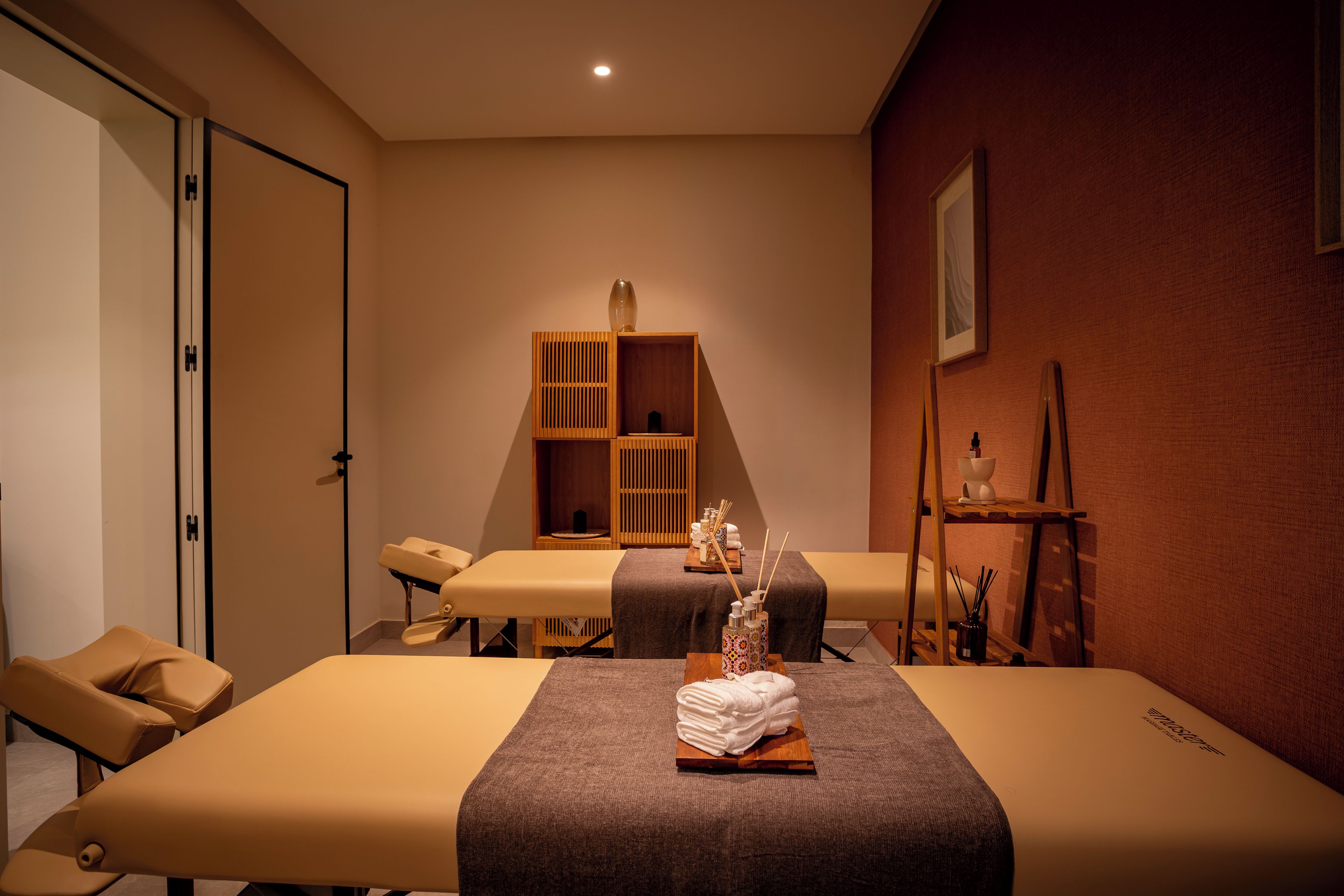 A massage therapy room with two massage tables, a wooden cabinet with shelves, and a small shelf with a vase and candles.