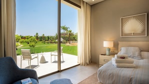 Habitación de hotel con un ventanal grande, una cama, una silla, una mesita de noche con lámpara y vista a un jardín.