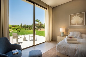 Une chambre d’hôtel avec une grande fenêtre, un lit, une chaise, une table de chevet avec une lampe et une vue sur un jardin.