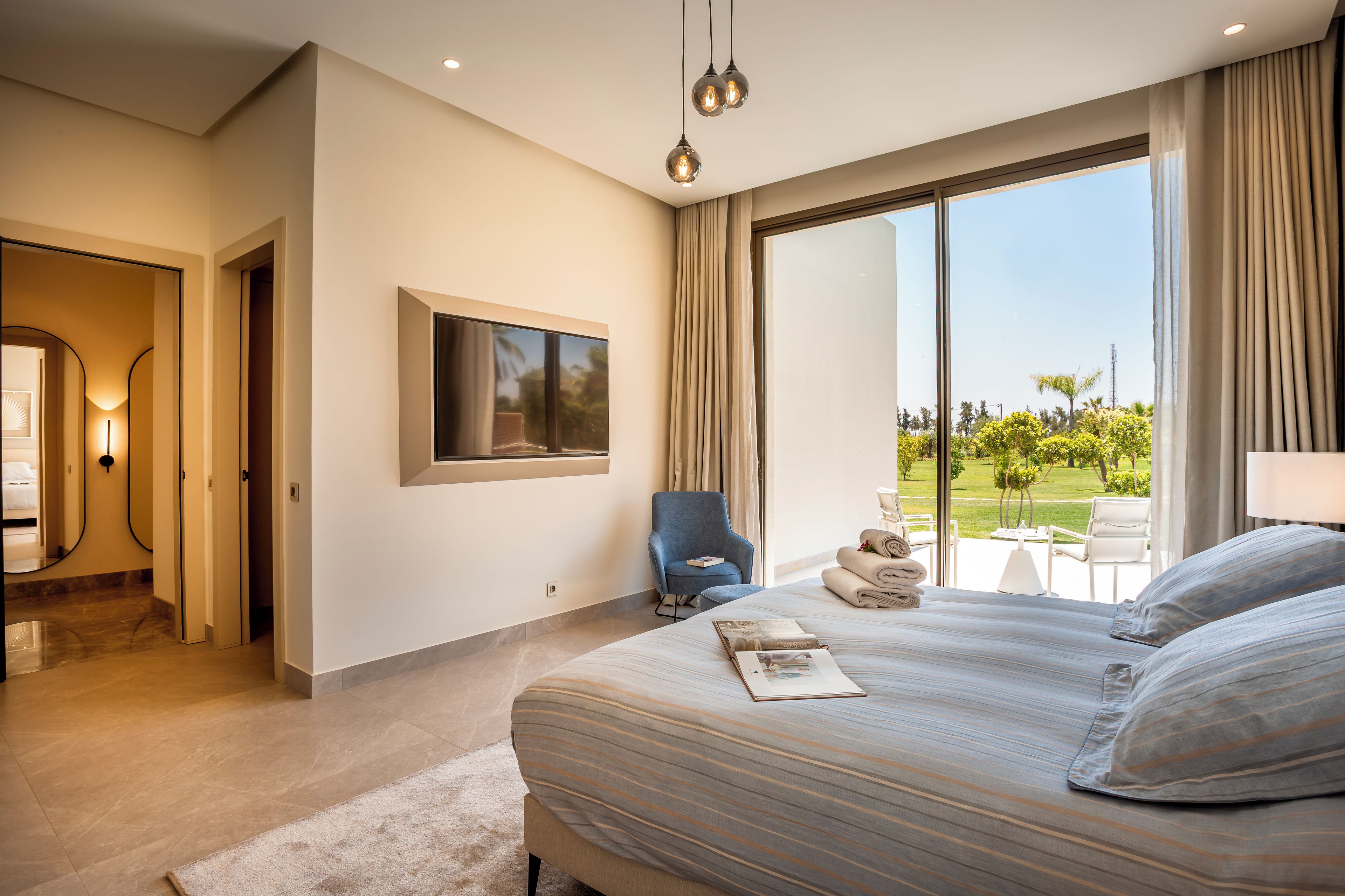 A modern hotel room with a large bed, a seating area with a chair and a table, and a view of a garden through the window.