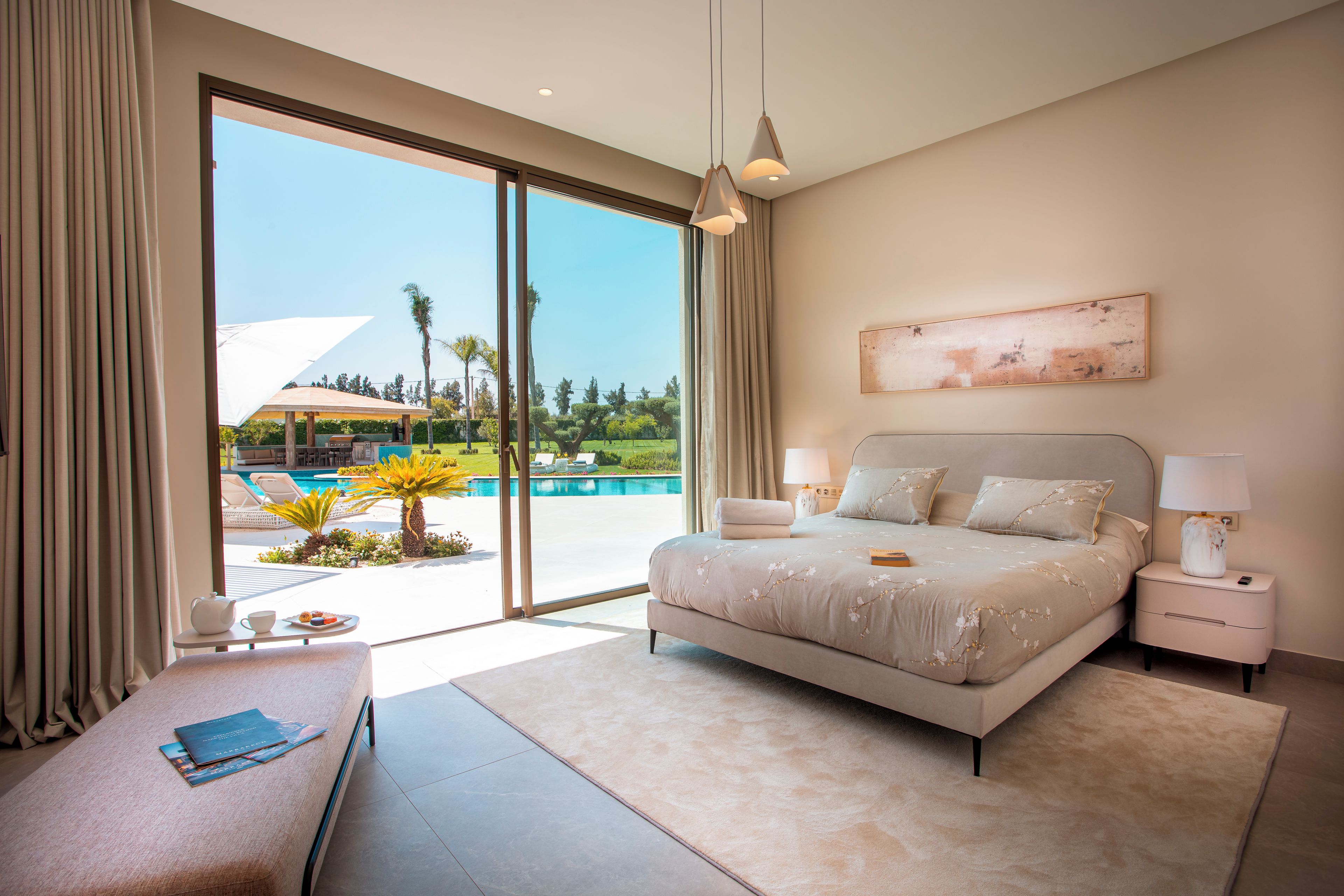 A bedroom with a large bed, a nightstand, a bedside table, a view of a swimming pool, and a door leading to an outdoor area.