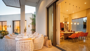 Un área de comedor al aire libre con mesas y sillas redondas de mimbre, una puerta de vidrio que da a un comedor y un interior moderno con iluminación colgante.