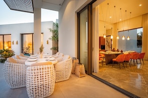 Un espace extérieur aménagé pour les repas, avec des tables et des chaises rondes en osier, une porte vitrée donnant sur une salle à manger, et un intérieur moderne agrémenté de luminaires suspendus.