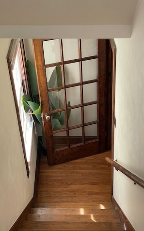 Eine Holztreppe mit Handlauf, die zu einem Raum mit einem großen Fenster und einer Tür mit Gittermuster führt.