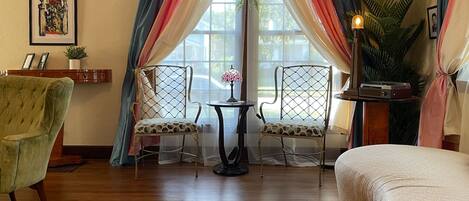 A room with a wooden floor, two chairs, a small table, a lamp, and a bed.