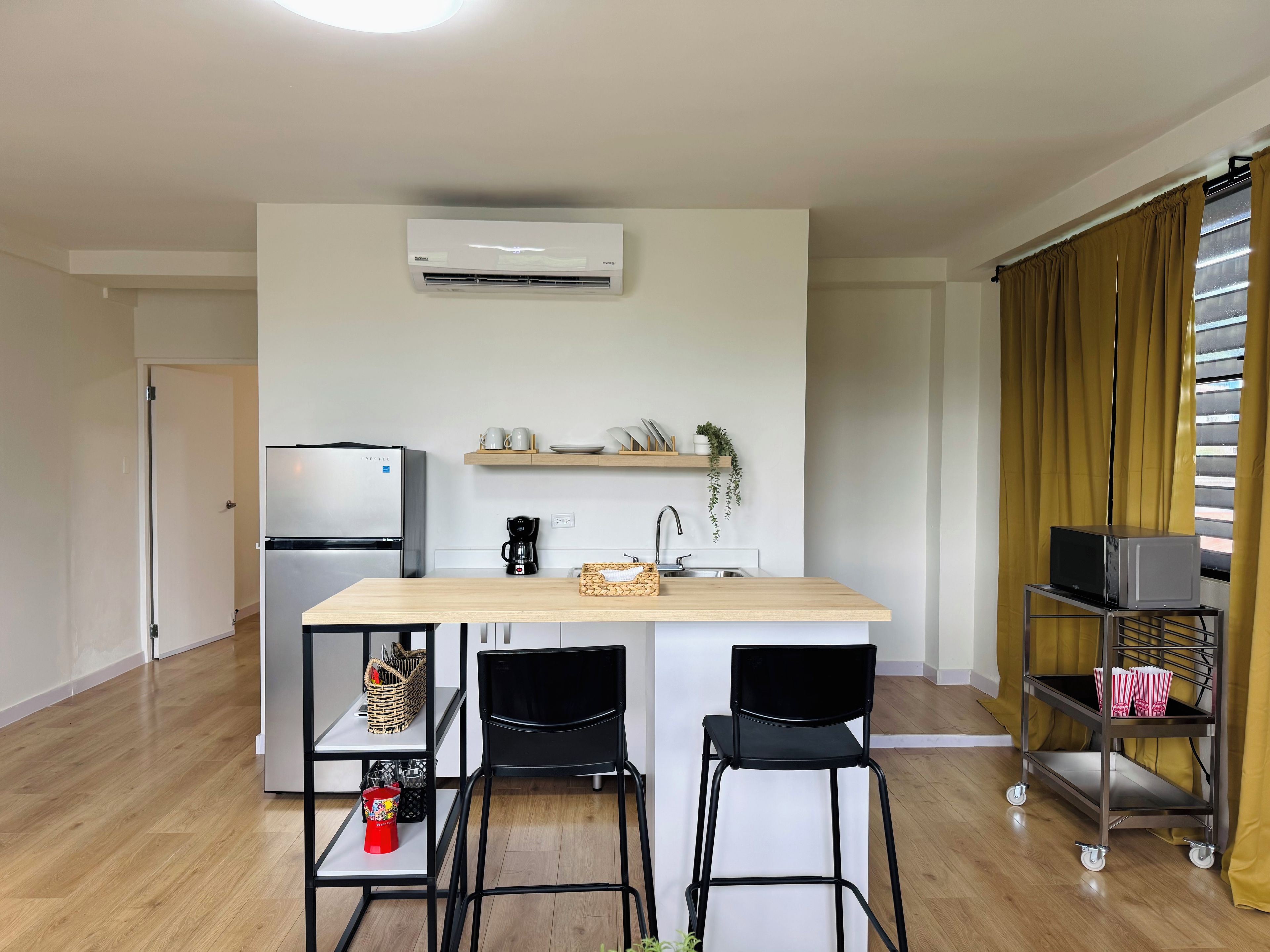 Cocina compacta con refrigerador integrado, cafetera y microondas en un estante. Hay dos sillas y una mesita en el centro.