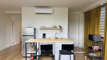 Cocina compacta con refrigerador integrado, cafetera y microondas en un estante. Hay dos sillas y una mesita en el centro.