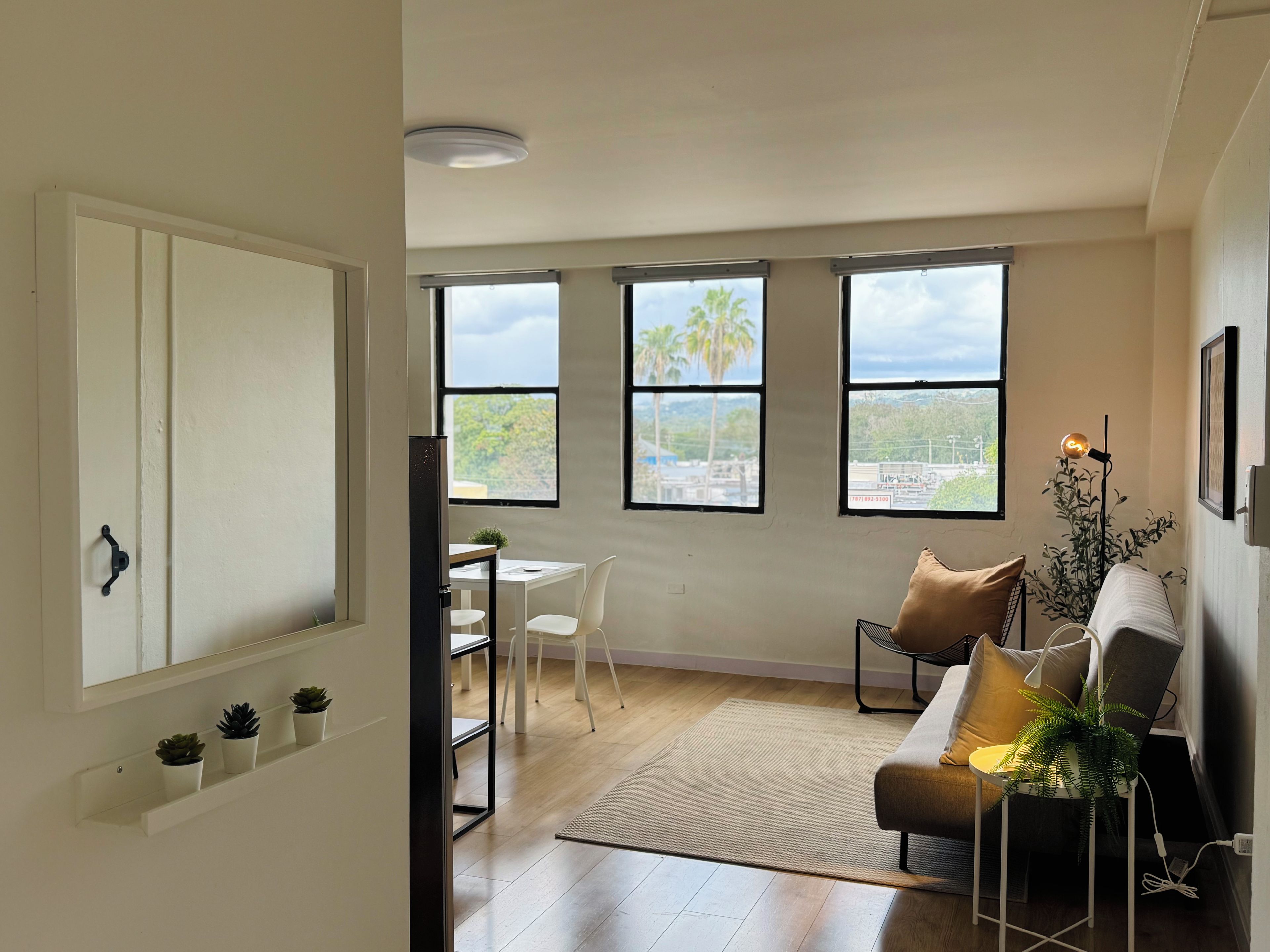 Una sala moderna con un sofá, una mesa de centro y un comedor con una mesa y sillas. Amplias ventanas ofrecen vistas a palmeras y edificios.
