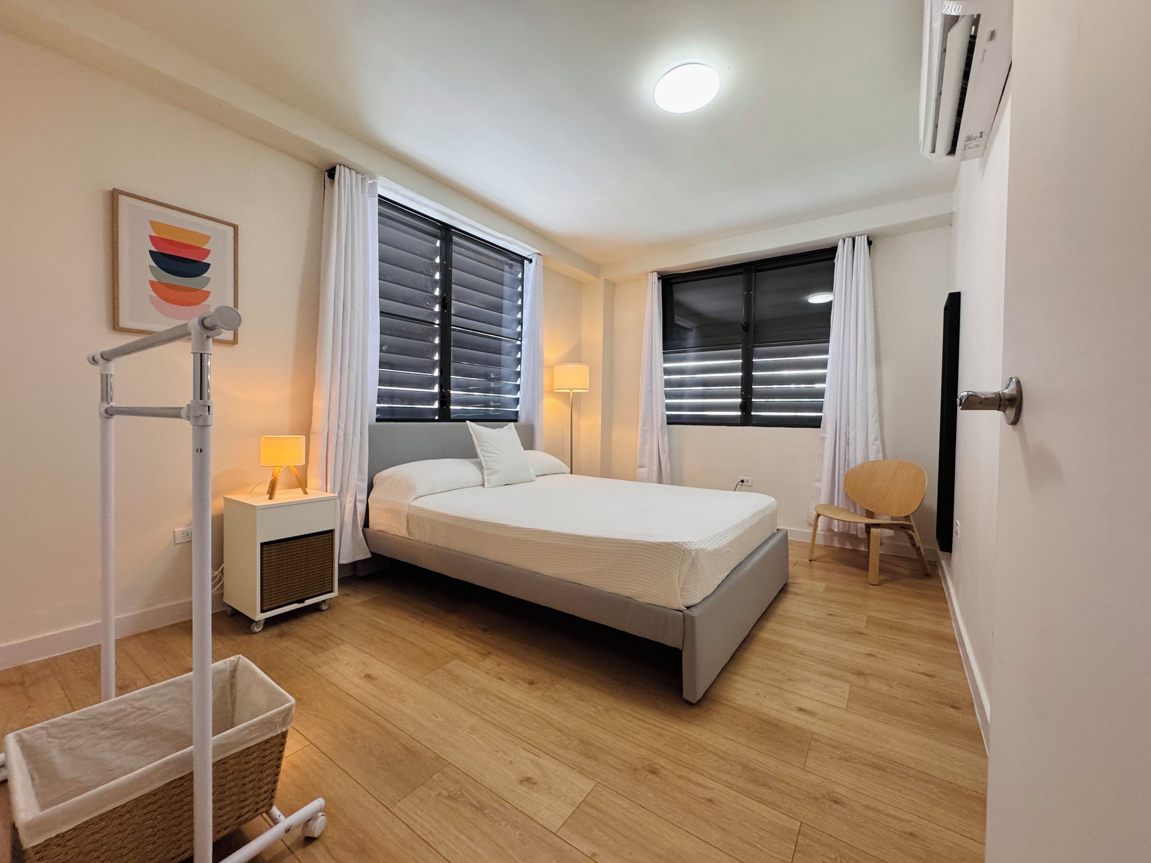 Quarto de hotel com cama, duas mesas de cabeceira, luminária, cadeira, abajur e ar-condicionado fixado na parede.