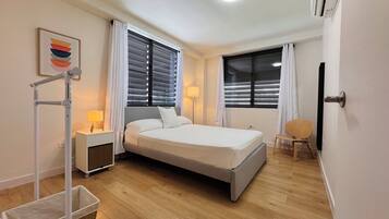 Quarto de hotel com cama, duas mesas de cabeceira, luminária, cadeira, abajur e ar-condicionado fixado na parede.
