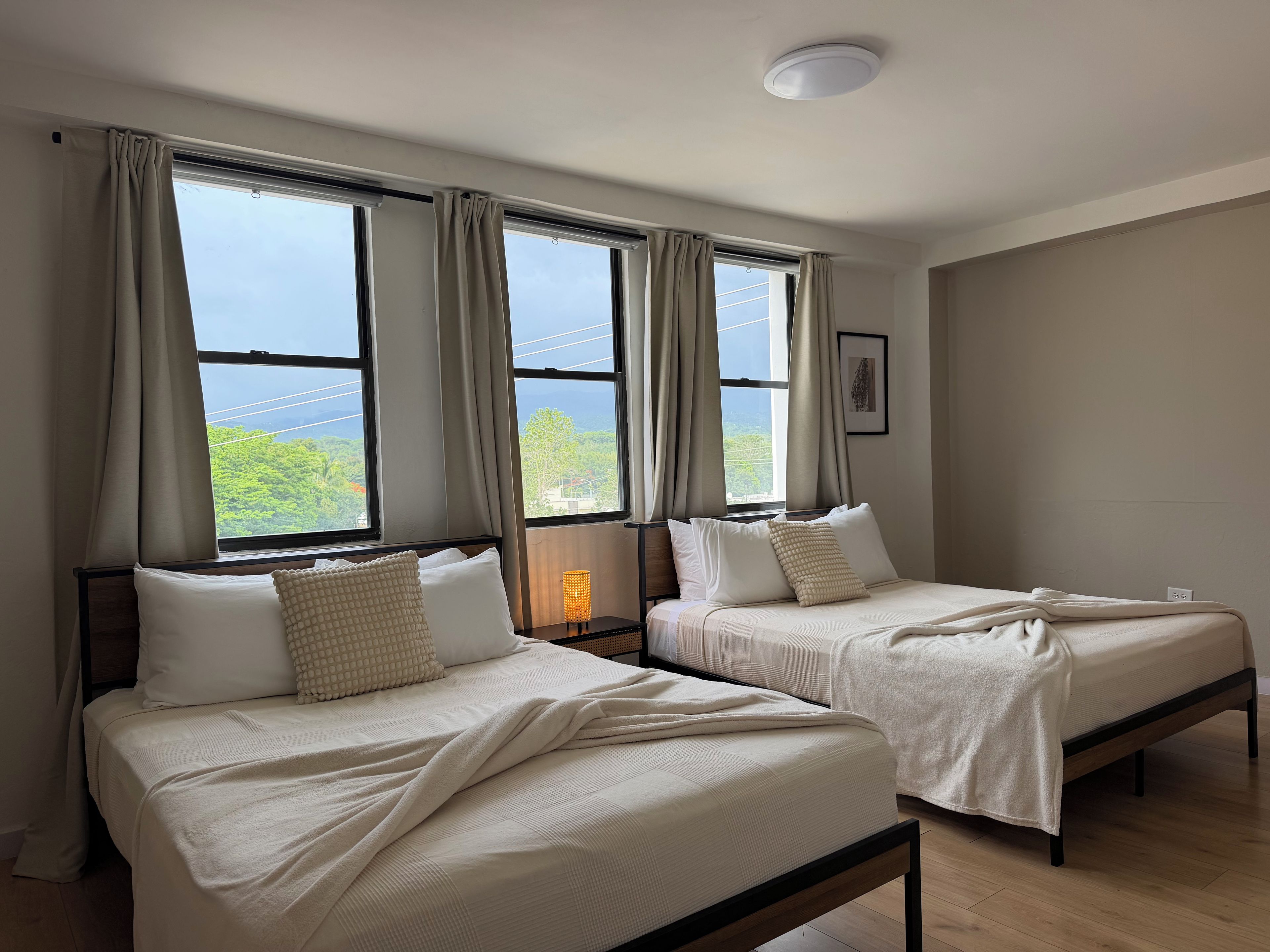 A room with two beds, large windows, a ceiling light, and a wall-mounted picture.