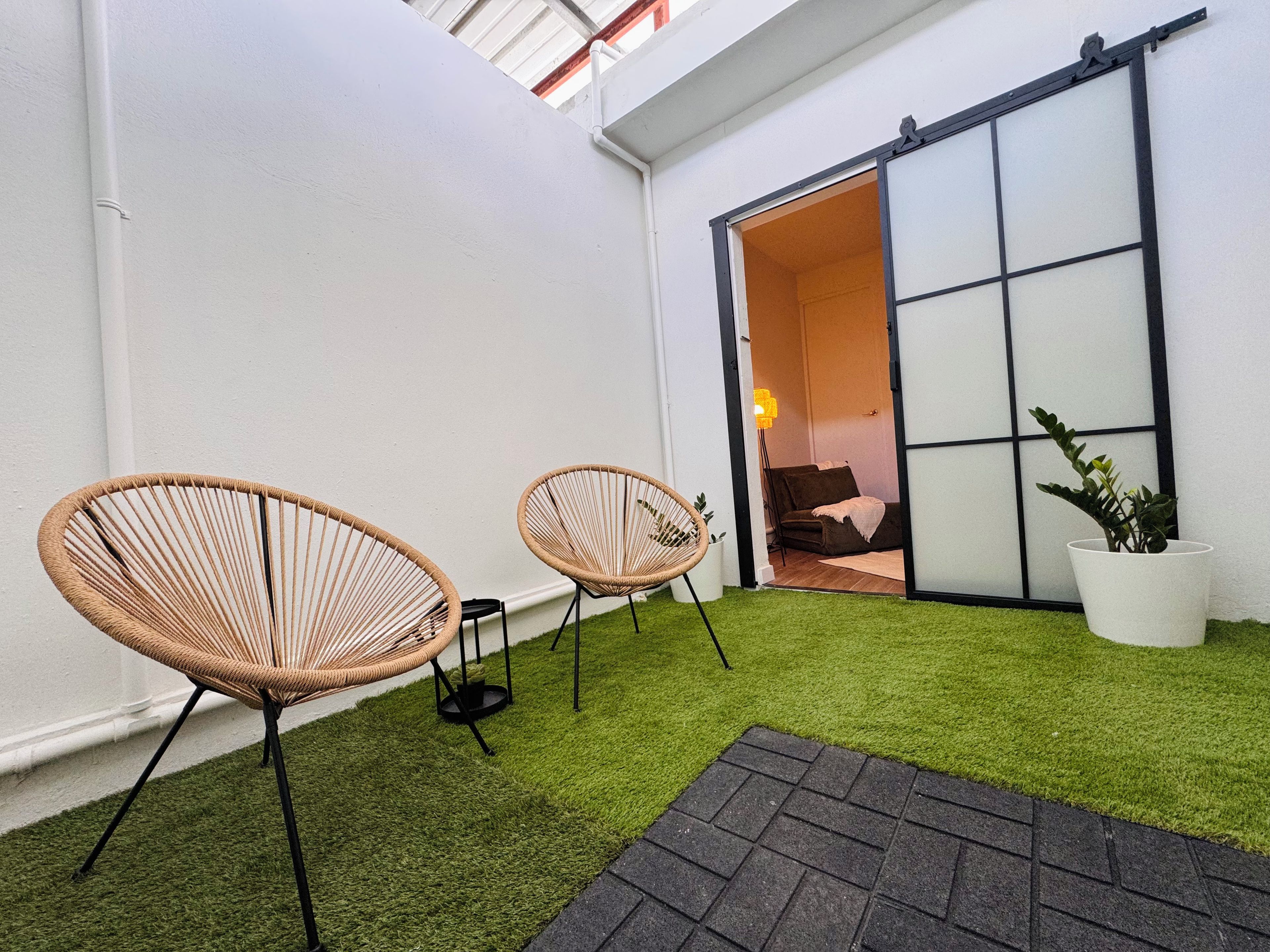 Un pequeño espacio exterior con césped artificial, dos sillas de mimbre, una planta en maceta y una puerta corredera que da a un espacio interior.