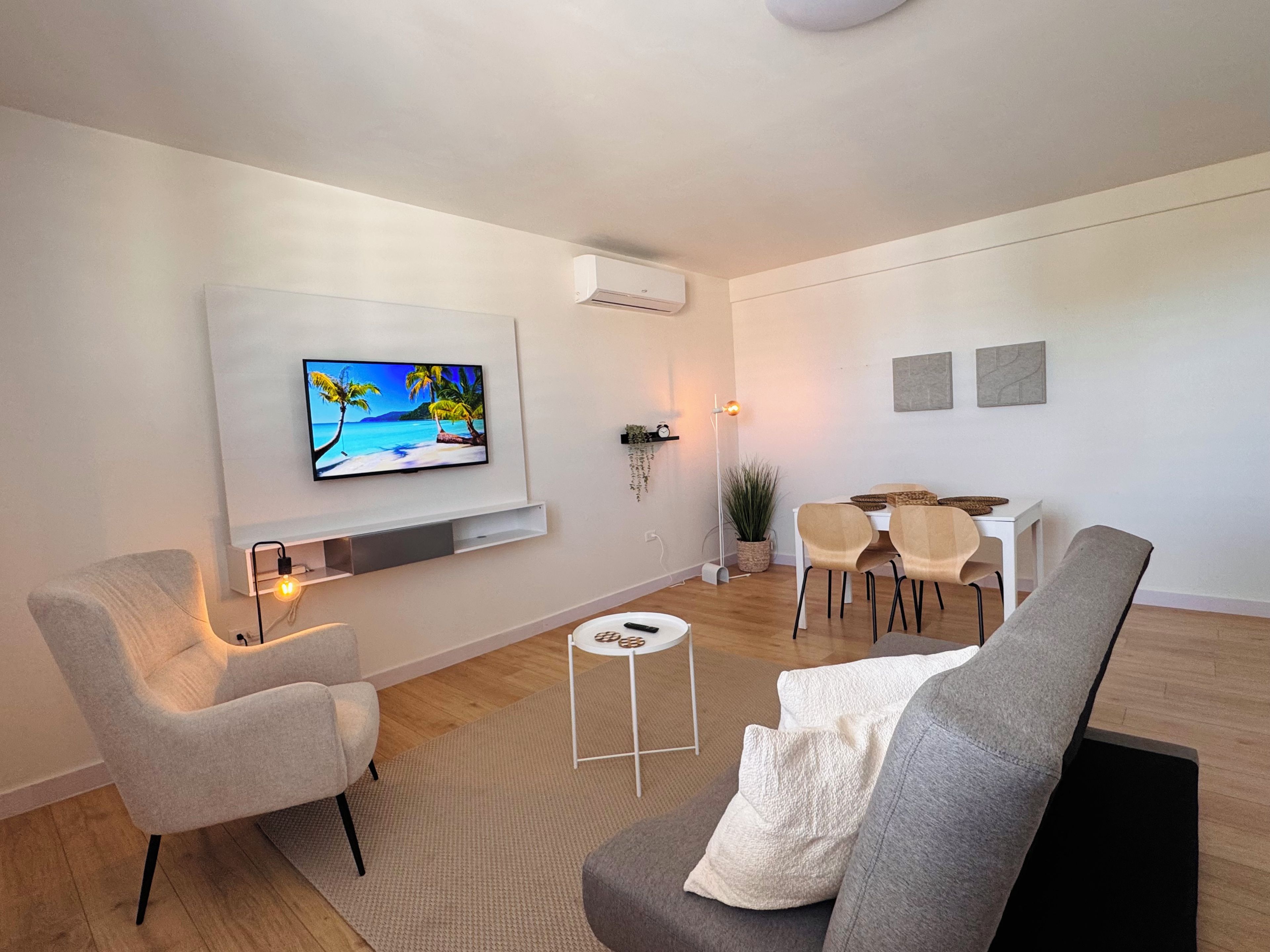 Een moderne woonkamer met een flatscreen-televisie, een eethoek met een witte tafel en stoelen, en een grijze bank.