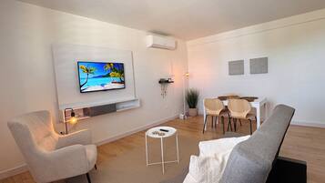 Een moderne woonkamer met een flatscreen-televisie, een eethoek met een witte tafel en stoelen, en een grijze bank.