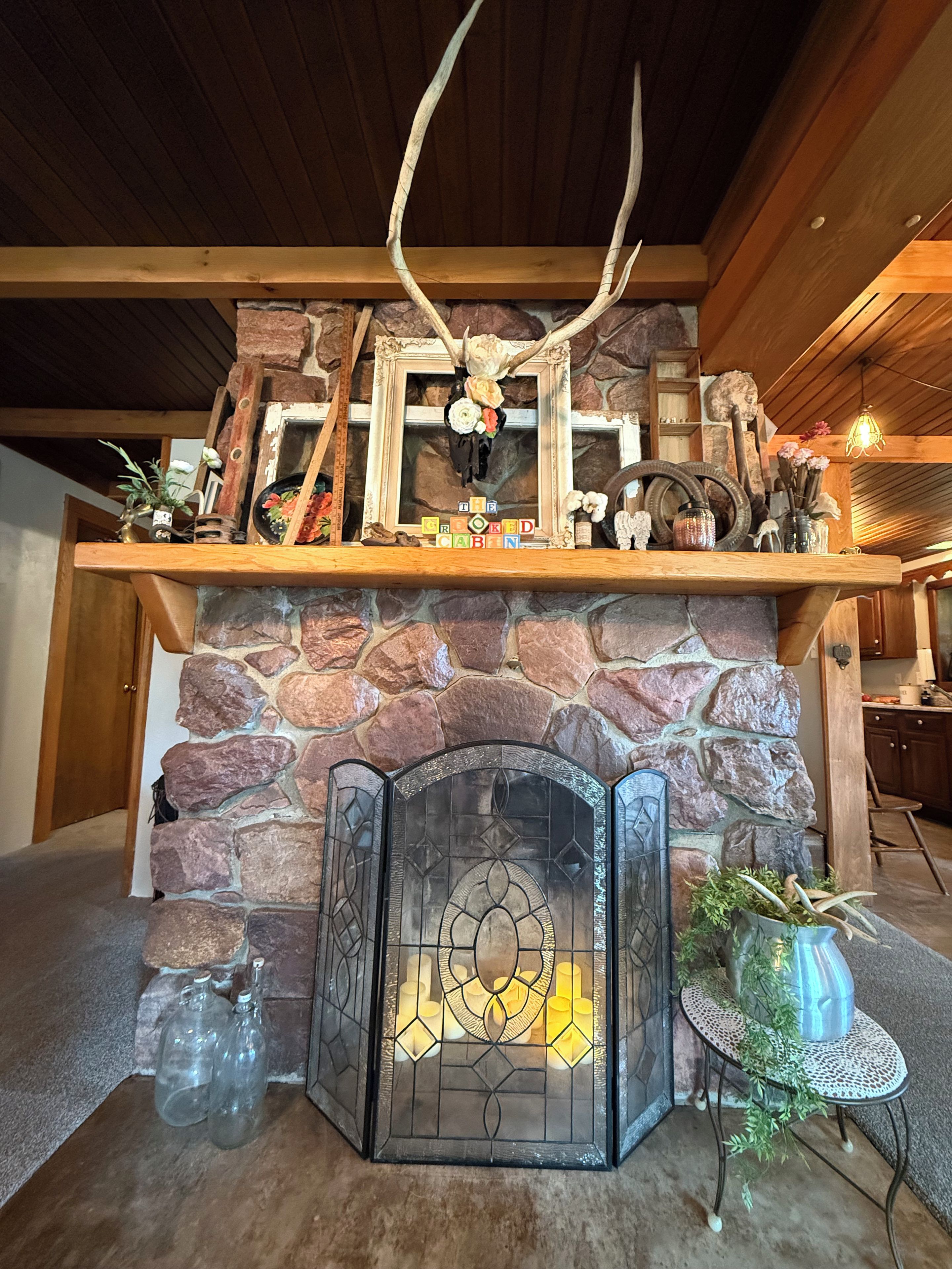 Una chimenea de piedra con una puerta de vitral, un fuste de madera adornado con astas y flores, y una pequeña mesa con plantas.