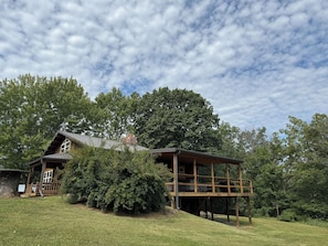 A wooden cabin with a deck, surrounded by trees and a grassy hill.
