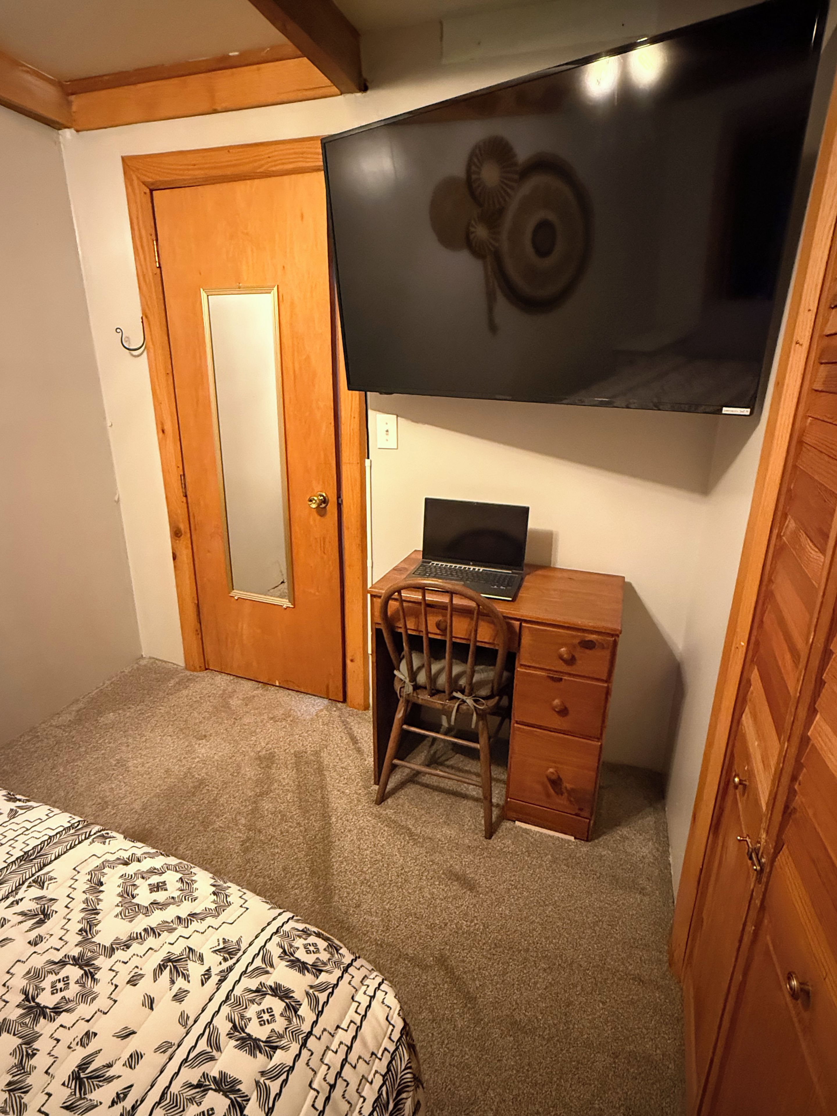 Un dormitorio con cama, escritorio con una computadora portátil, silla, armario de madera y televisor montado en la pared.