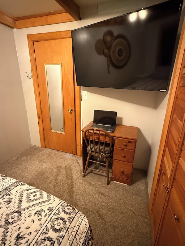 A bedroom with a bed, a desk with a laptop, a chair, a wooden wardrobe, and a television mounted on the wall.