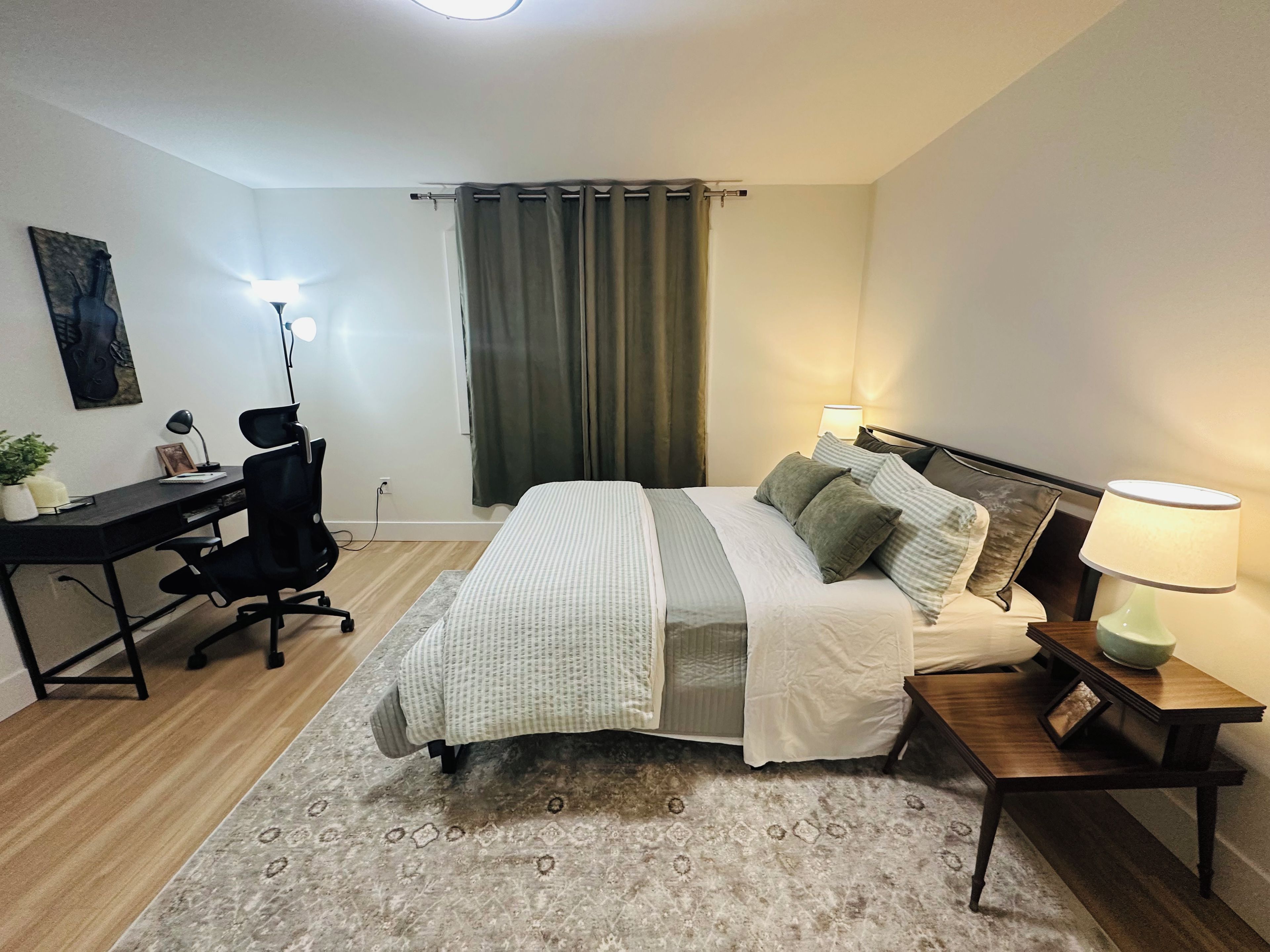 A hotel room with a bed, desk, chair, lamp, and a painting on the wall.