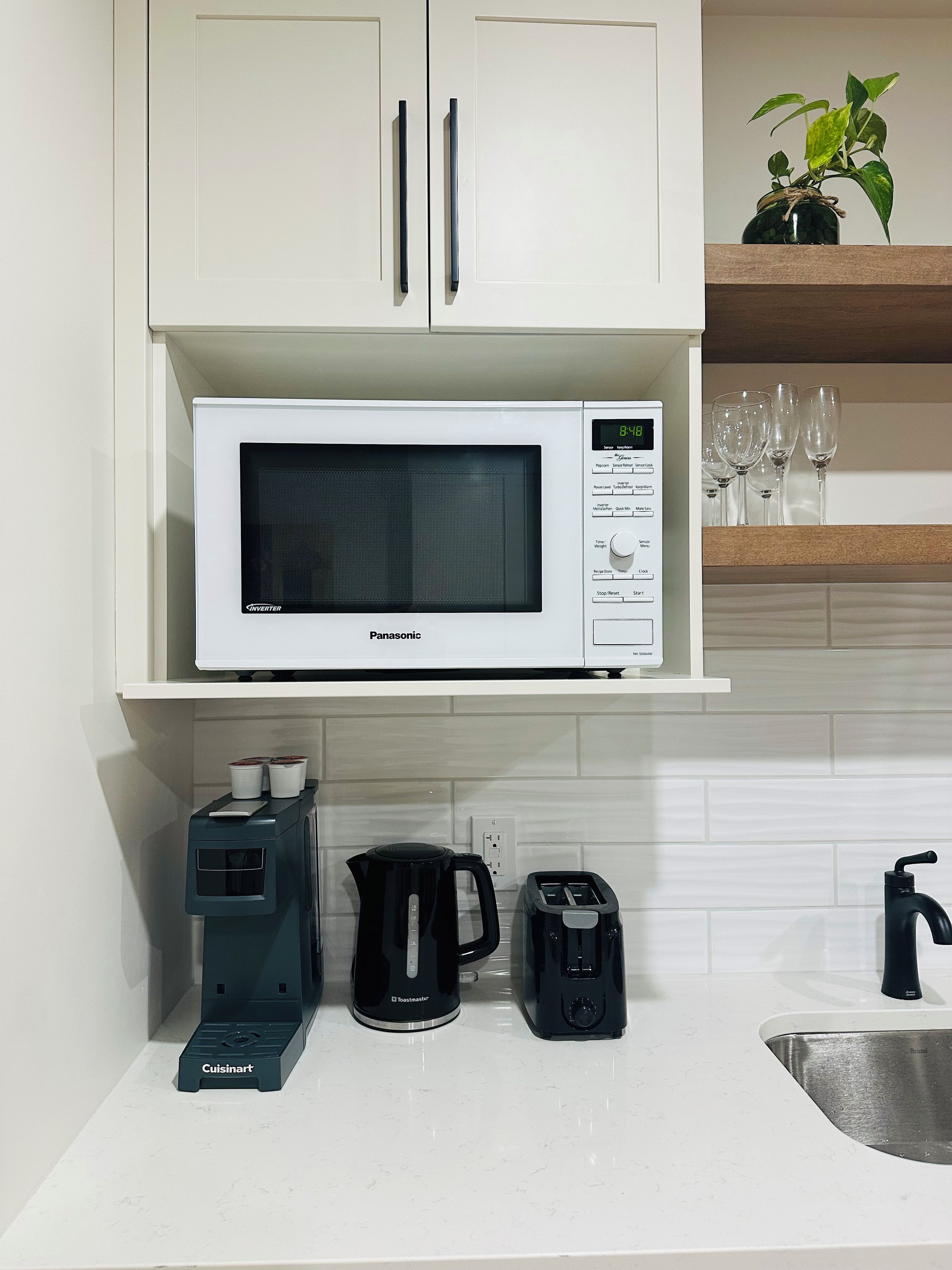 Une cuisine équipée d’un four à micro-ondes, d’un grille-pain et d’une machine à café sur le plan de travail, et d’une plante posée sur l’étagère au-dessus.
