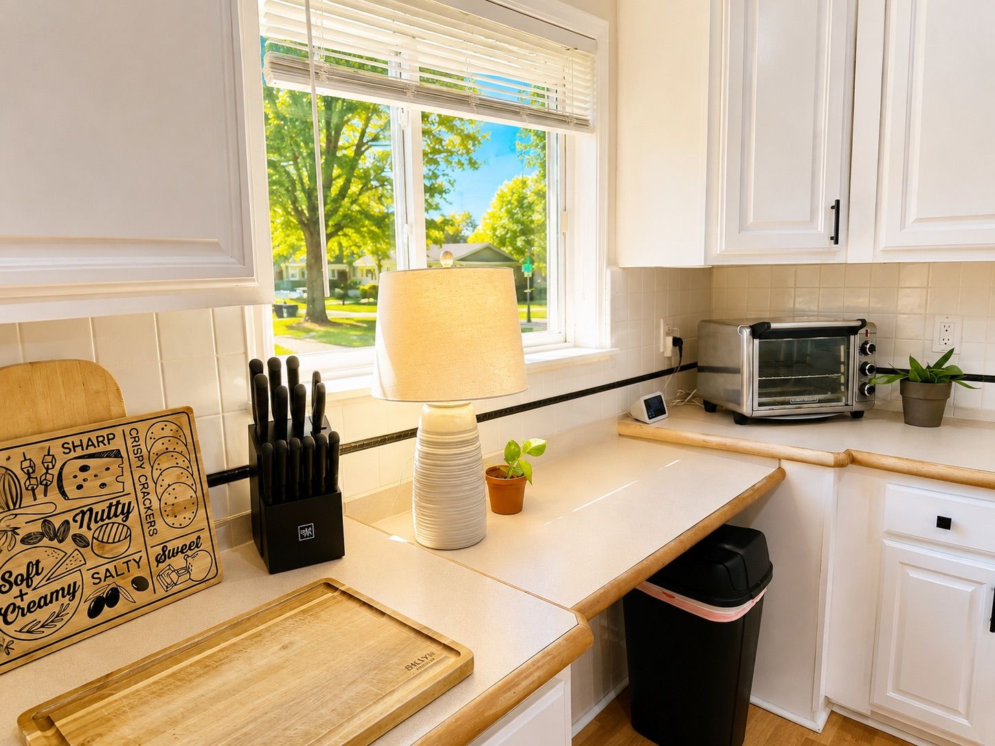 Une cuisine avec des meubles blancs, un grille-pain, une fenêtre avec des stores, une lampe, une poubelle et une planche à découper.