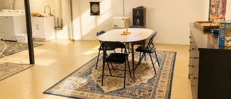 Una habitación con una mesa de comedor, sillas, un tapete estampado, una lavadora, un armario y un letrero de neón.