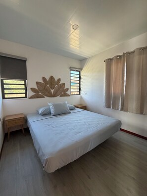 A bedroom with a bed, two nightstands, a window with blinds, a wall-mounted light fixture, and a curtain.