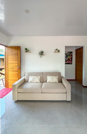 A neutral-toned living room with a beige sofa, two patterned cushions, and three wall-mounted shelves with potted plants. - FARE HANI - Combining comfort and privacy with a pool (Maatea)