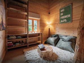 Ein gemütliches Blockhaus-Interieur mit einem hölzernen Bücherregal, einem Fenster mit Blick auf Grünflächen, einer Lampe und einem Sofa mit Kissen.