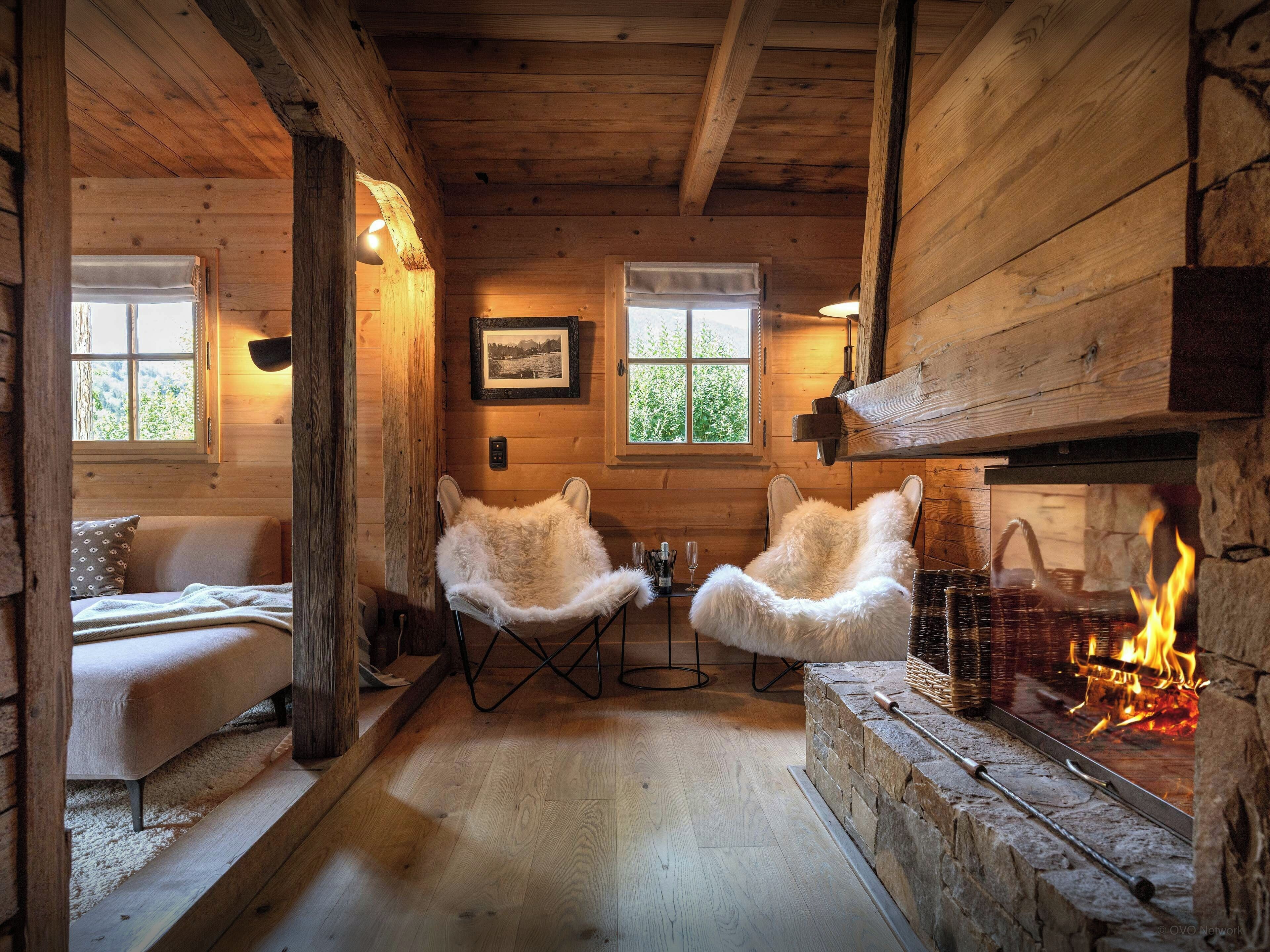 Interior acogedor de una cabaña con chimenea de piedra, dos sillones cubiertos de piel, un sofá y un ventanal con vistas al follaje.