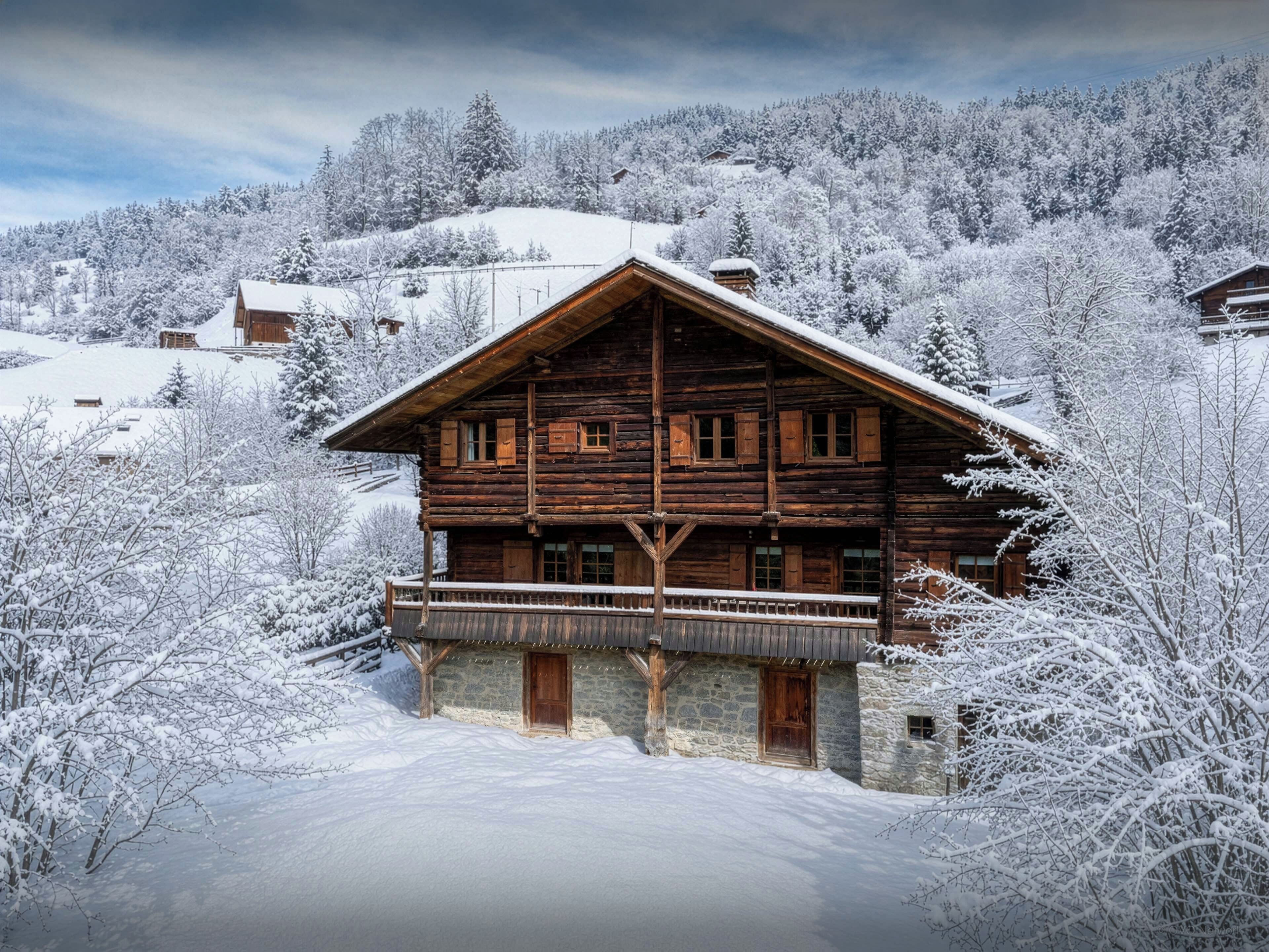 A wooden log cabin with a balcony, surrounded by snow-covered trees and a mountainous backdrop.