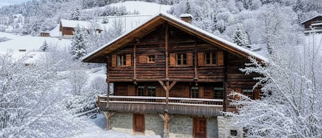 Una baita di legno con un balcone, immersa in un paesaggio innevato di alberi e montagne.