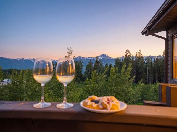 A balcony with two wine glasses and a plate of food, overlooking a mountainous landscape.