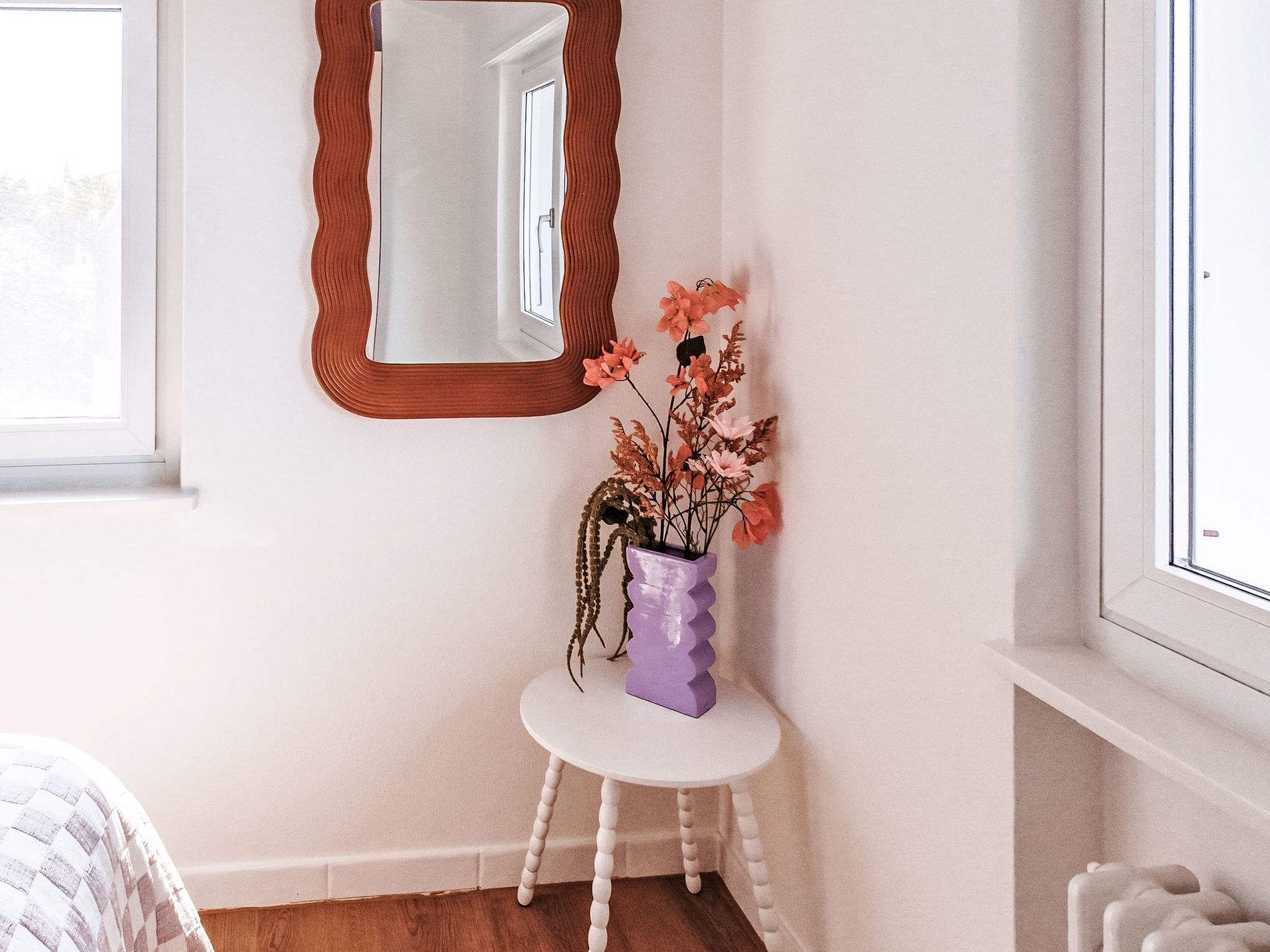 Une pièce avec un mur blanc, une table ronde blanche ornée d’un vase violet et un miroir à cadre ondulé.