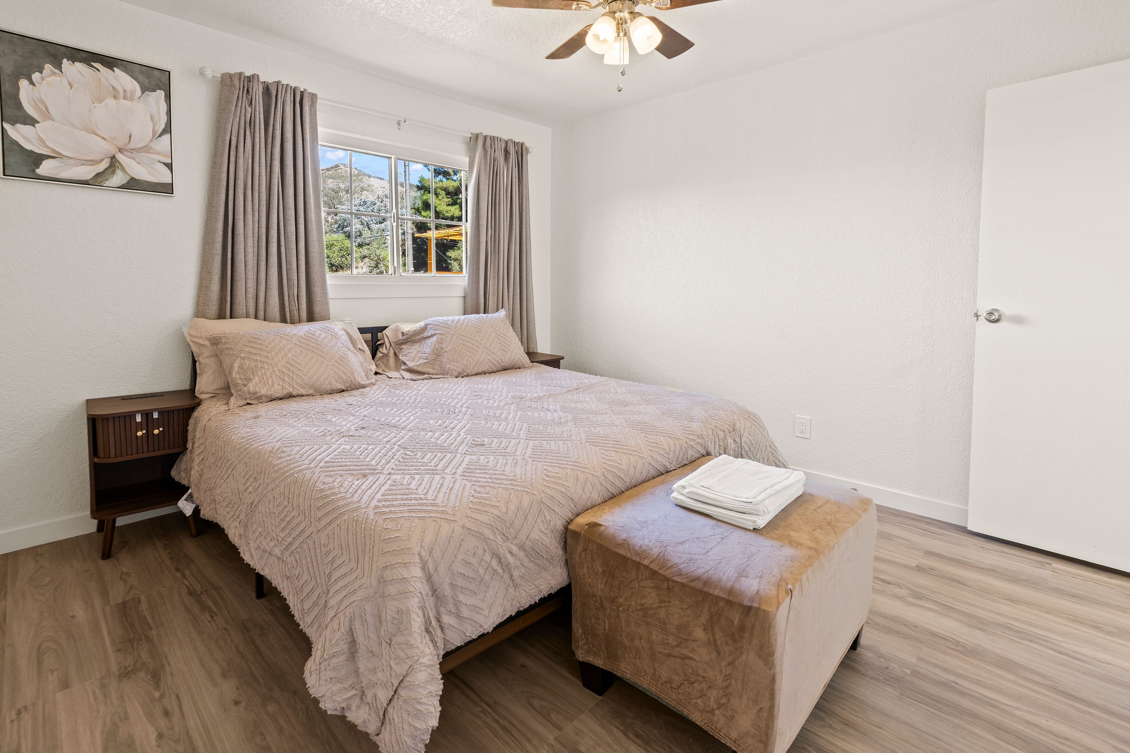 Une chambre à coucher comprenant un lit, une table de chevet, un banc, une fenêtre avec des rideaux et un ventilateur de plafond.