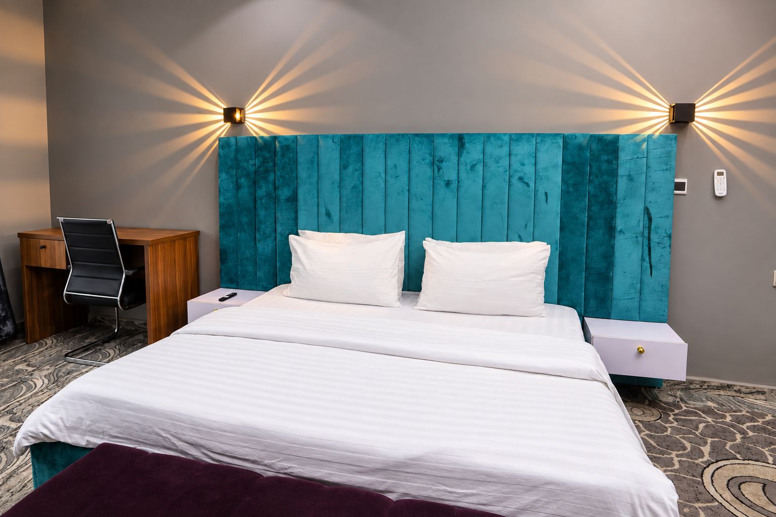 A hotel room with a bed, two bedside tables, a desk, a chair, and a wall-mounted light fixture.