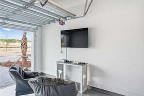 Ein Zimmer mit an der Wand montiertem Flachbildfernseher, einem weißen Konsolentisch mit einigen Gegenständen, einem grauen Sofa und einer Palme.