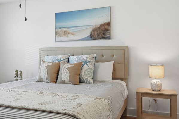 Ein Schlafzimmer mit einem Bett, einem Kopfteil, einem Nachttisch mit Lampe und einem an der Wand hängenden Bild von einem Strand.