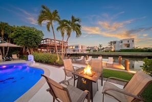 A poolside area with lounge chairs, a fire feature, and a view of a marina with buildings. - New! Seaside Sunset! Luxury Canal/Pool Home! 1 Mile to the Beach! (Naples)