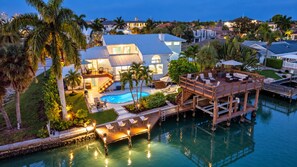 A waterfront property with a swimming pool, a deck, and a house with a large window overlooking the water. - New! Seaside Sunset! Luxury Canal/Pool Home! 1 Mile to the Beach! (Naples)