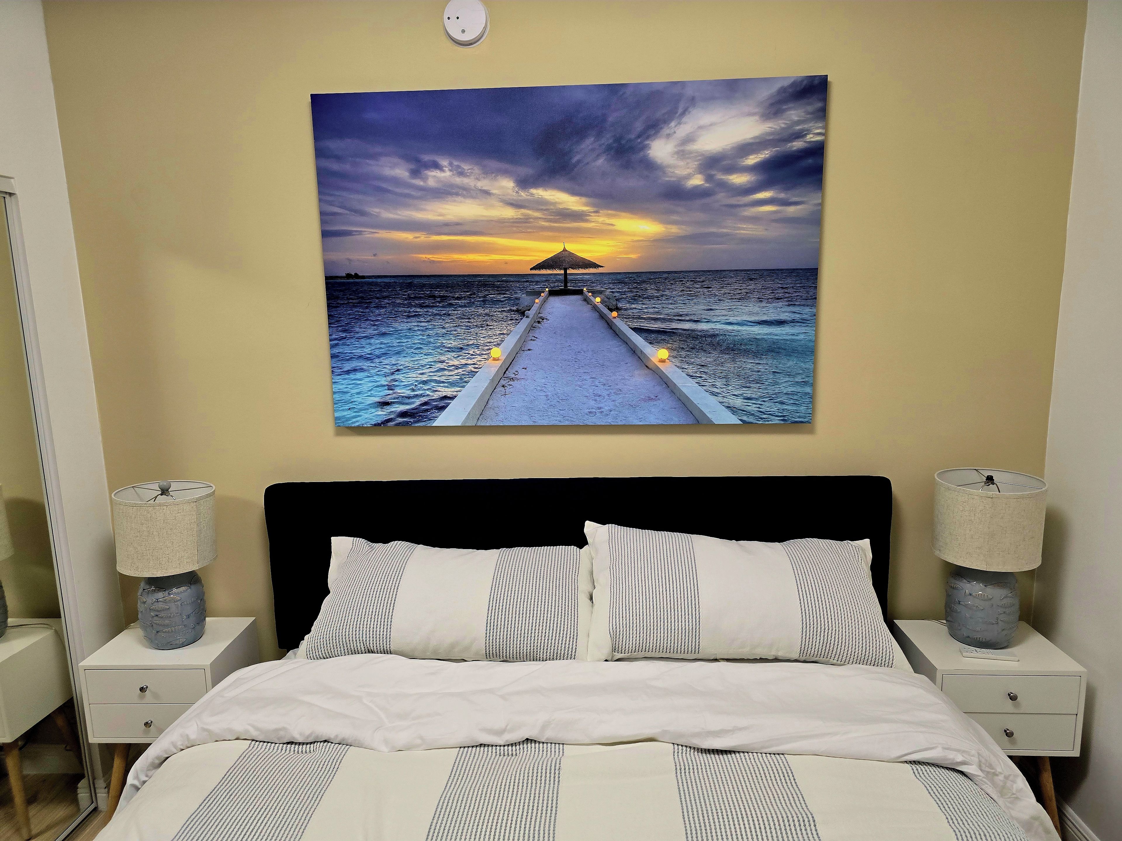 Un dormitorio con una cama, dos lámparas de noche, una mesita de noche y un cuadro colgado en la pared que muestra un atardecer sobre el océano.