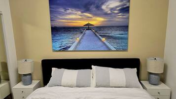 Un dormitorio con una cama, dos lámparas de noche, una mesita de noche y un cuadro colgado en la pared que muestra un atardecer sobre el océano.