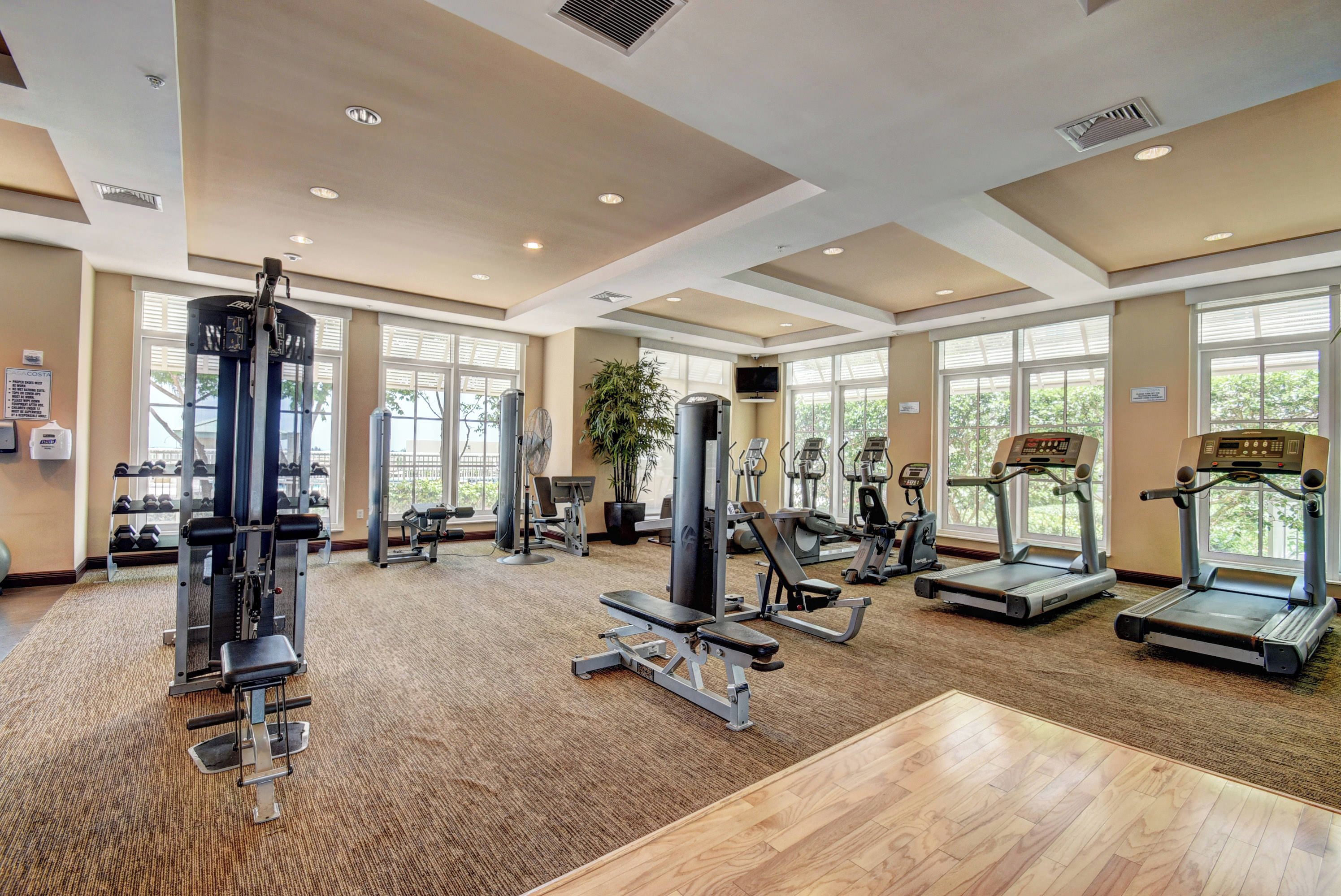 Un gimnasio espacioso con diversos equipos de ejercicio, ventanas amplias y suelo de madera.