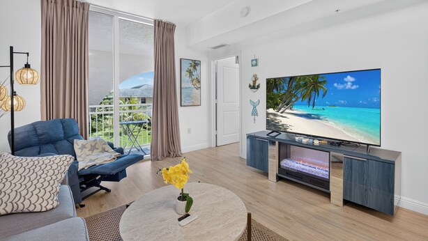 Ein modernes Wohnzimmer mit einem großen Flachbildfernseher, einem blauen Sofa, einem Couchtisch mit einer Pflanze und einem Balkon mit Vorhängen.