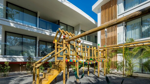 Een speeltuin met een houten klimstructuur en schommels, voor een modern gebouw met grote ramen.