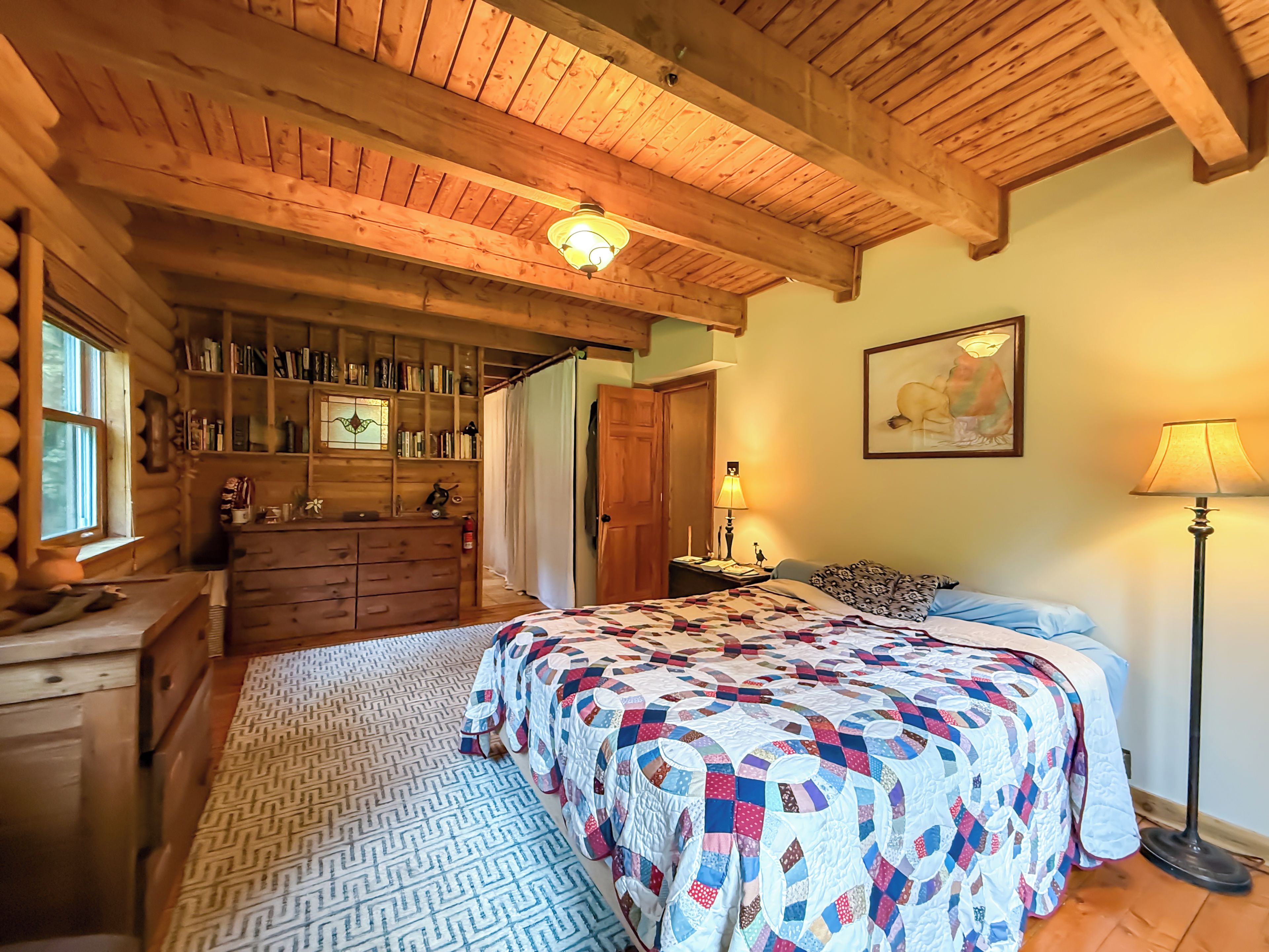 A log cabin bedroom with a large bed, wooden dresser, bookshelves, and a ceiling light.